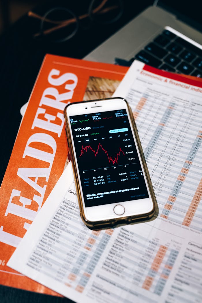 Smartphone displaying BTC chart on financial papers and laptop.