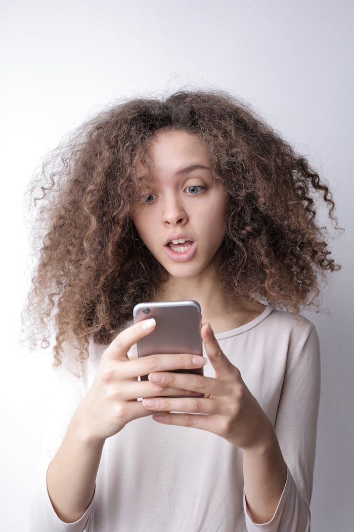 A young woman with curly hair looks surprised while reading something on her smartphone indoors.